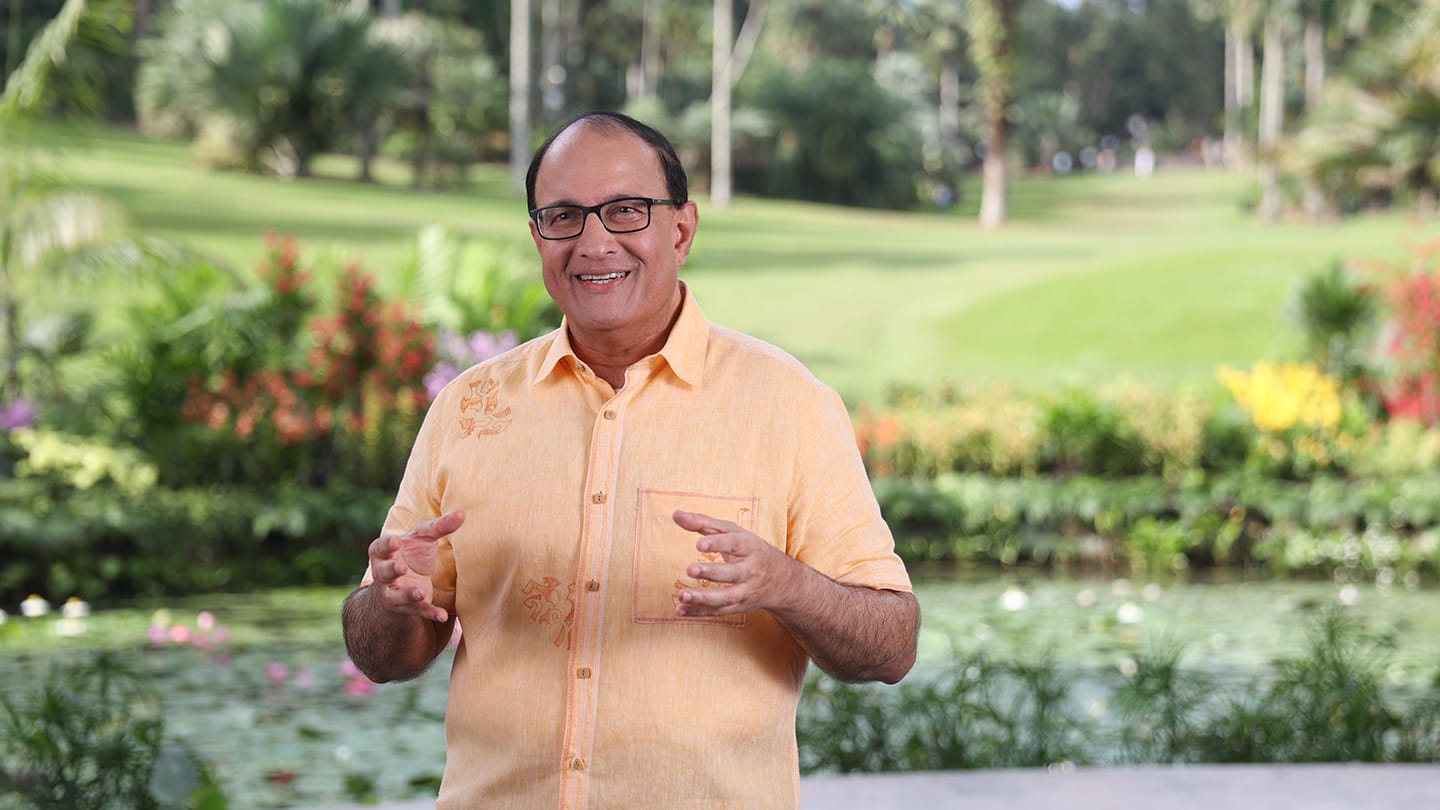 Man in orange shirt with glasses stands in garden setting, gesturing with hands.